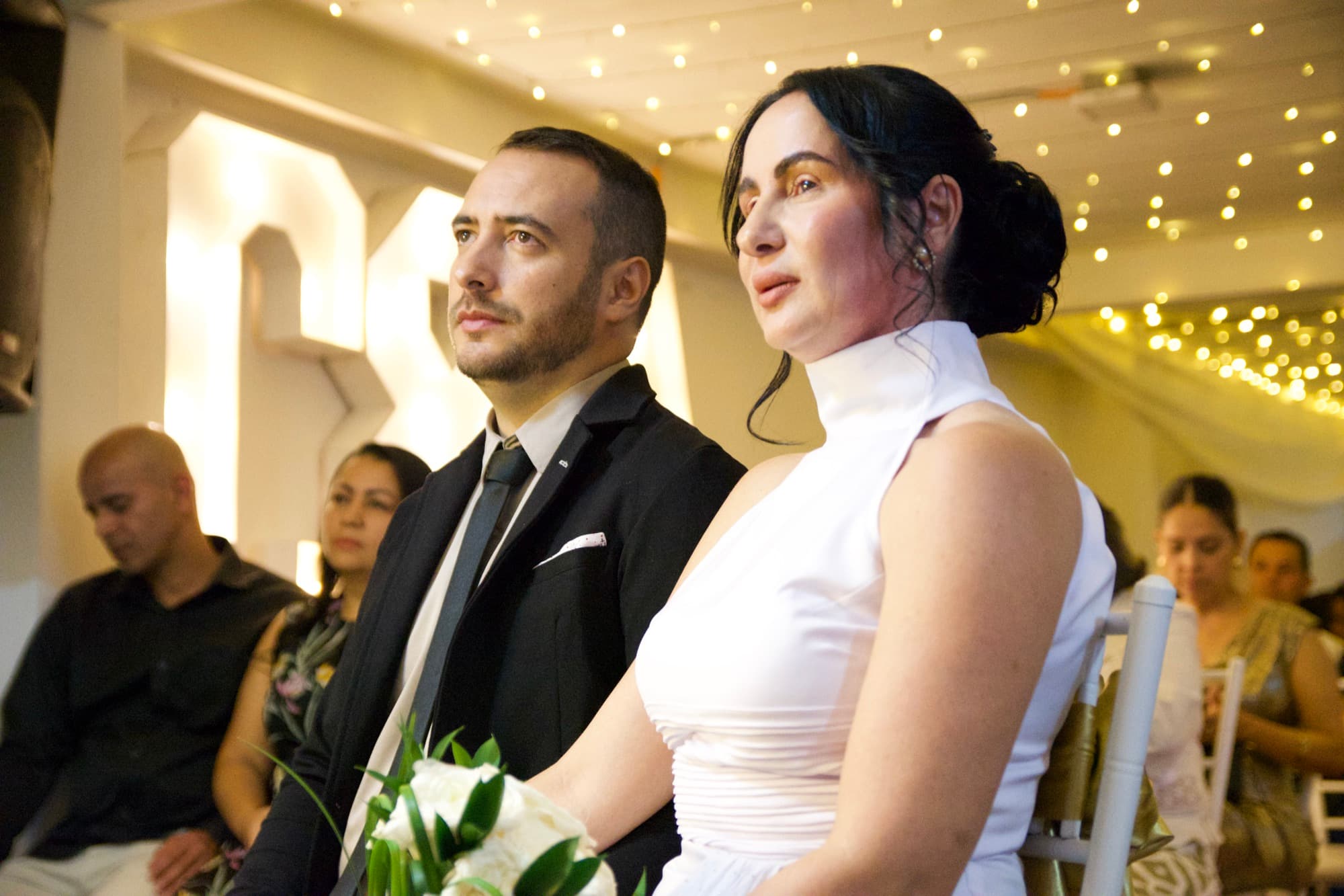 Los novios sentados de perfil escuchando la ceremonia, con los invitados detrás