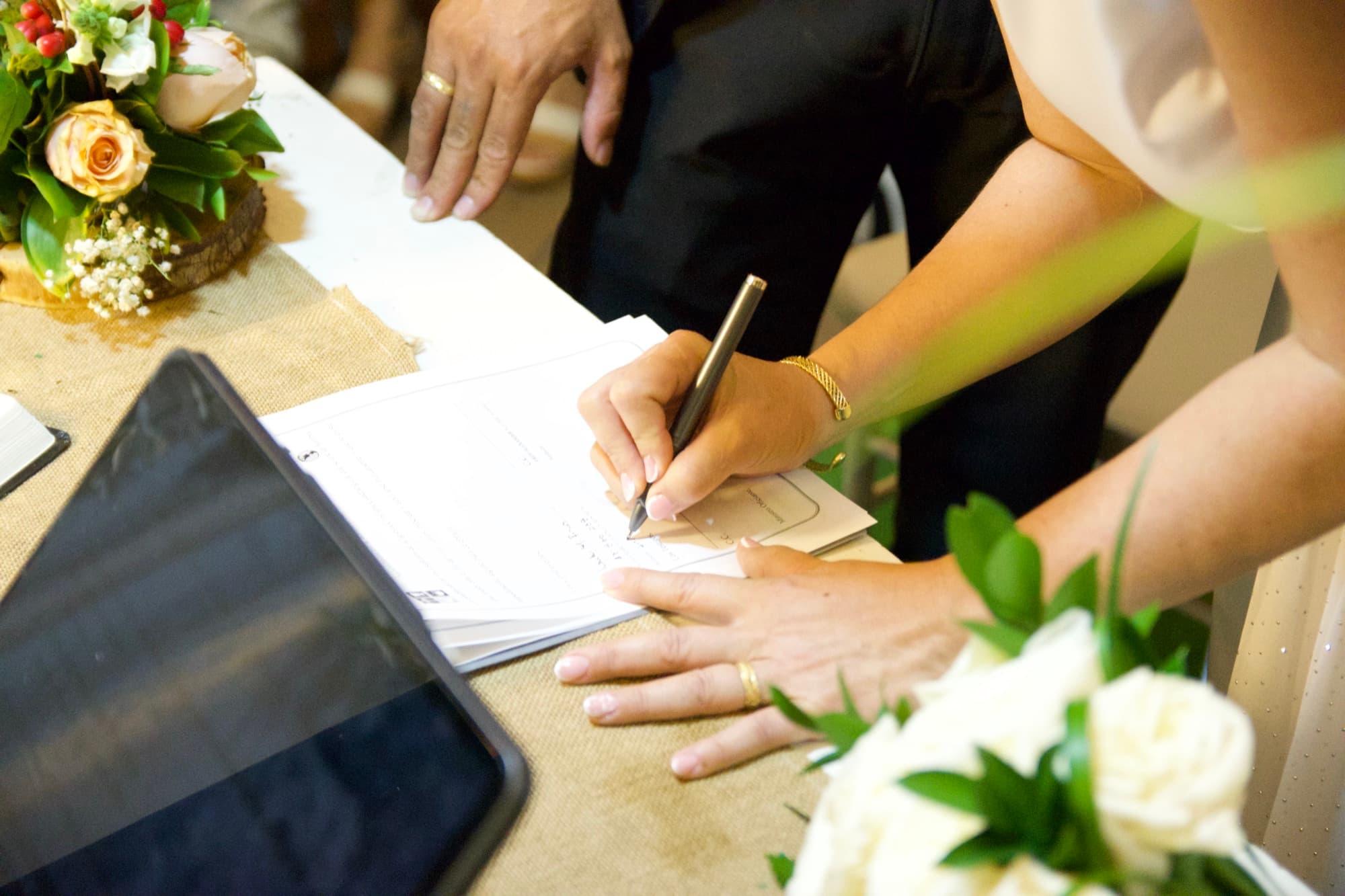 Manos firmando el acta matrimonial sobre la mesa, junto al ramo